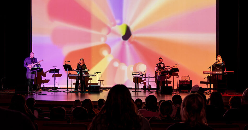 Photo of four musicians on stage with a colorful digital backdrop playing guitar, bass, keyboards with a crowd in the foreground.