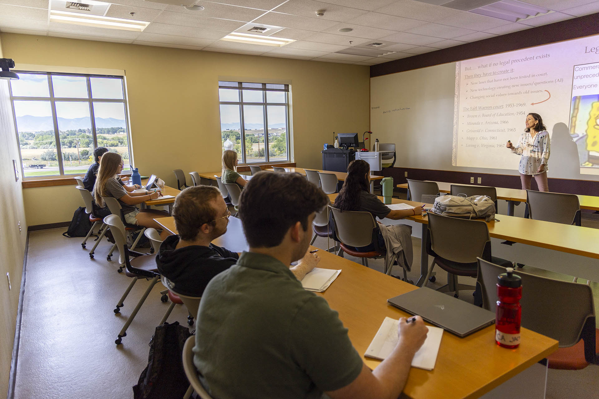 Students at desks in a classroom with a criminal justice instructor lecturing and mountains visible out the windows.