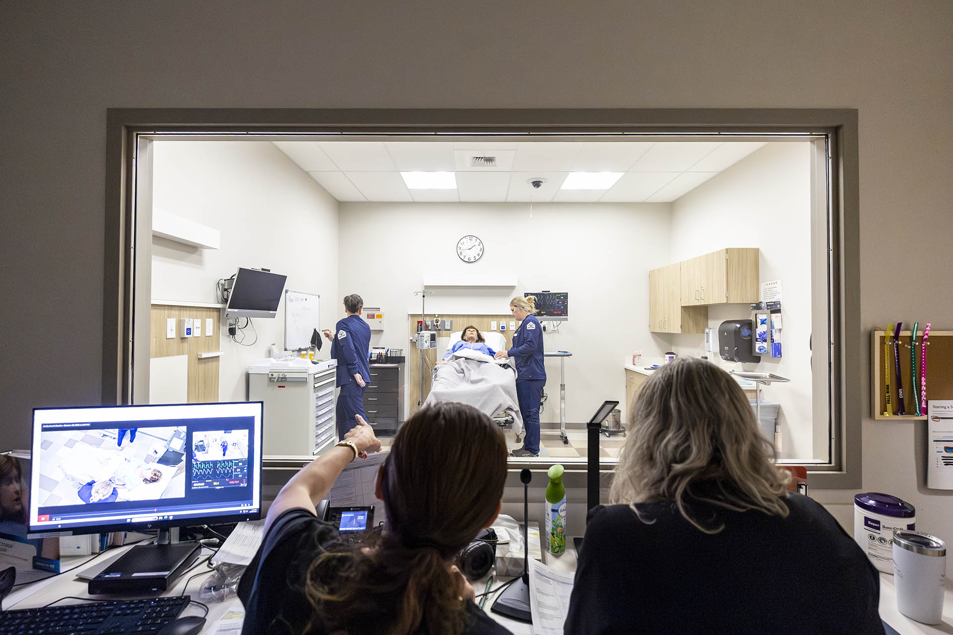Two instructors watch and guide a nursing simulation for second-year nursing students in a hi-fi simulation room