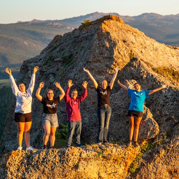 Sheridan College students smiling and holding arms upward on a large rock in the Bighorn Mountains.