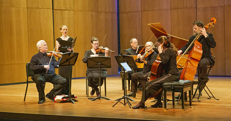 Wyoming Baroque performing on stage in Kinnison Hall with a harpsichord, upright bass, cello, viola, two violins, and a singer.
