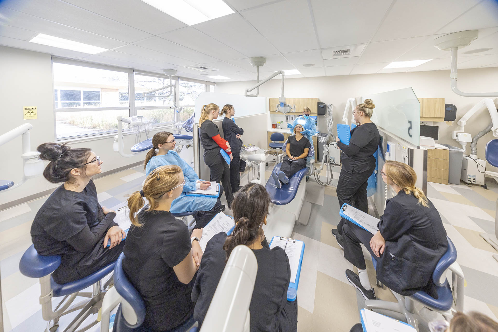 A group of Sheridan College Dental Hygiene students surrounding a dental chair as the instructor demonstrates a technique.