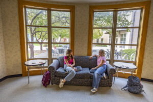 Two Sheridan College students sitting on a couch in front of two big paned windows with trees and bikes visible outside.