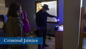 Criminal Justice student shines a purple light to reveal evidence during a mock investigation of a crime scene in a bedroom setting with the blinds drawn.