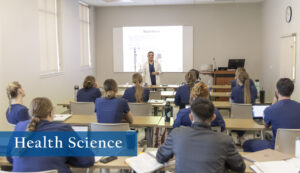Classroom full of nursing students as instructor stands in front of projection at the head of the classroom.