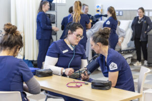 Nursing students checking blood pressure on each other, with a group of additional students in the background working with a hi fidelity maniken.