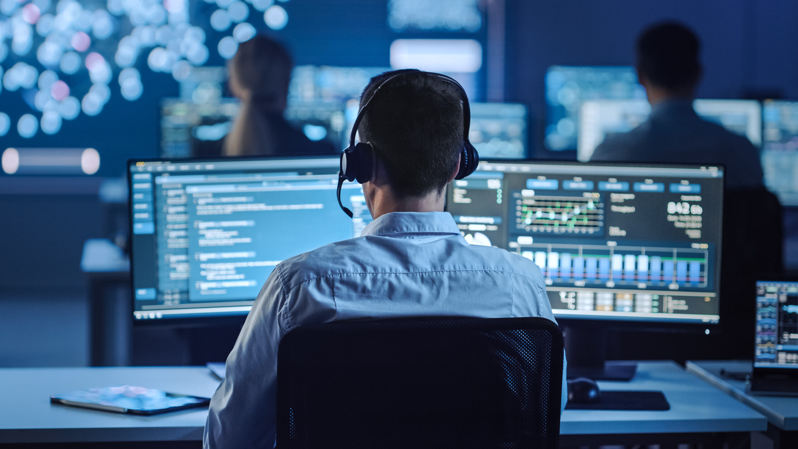 Cyber security staff member on looks two screens with colleagues in room