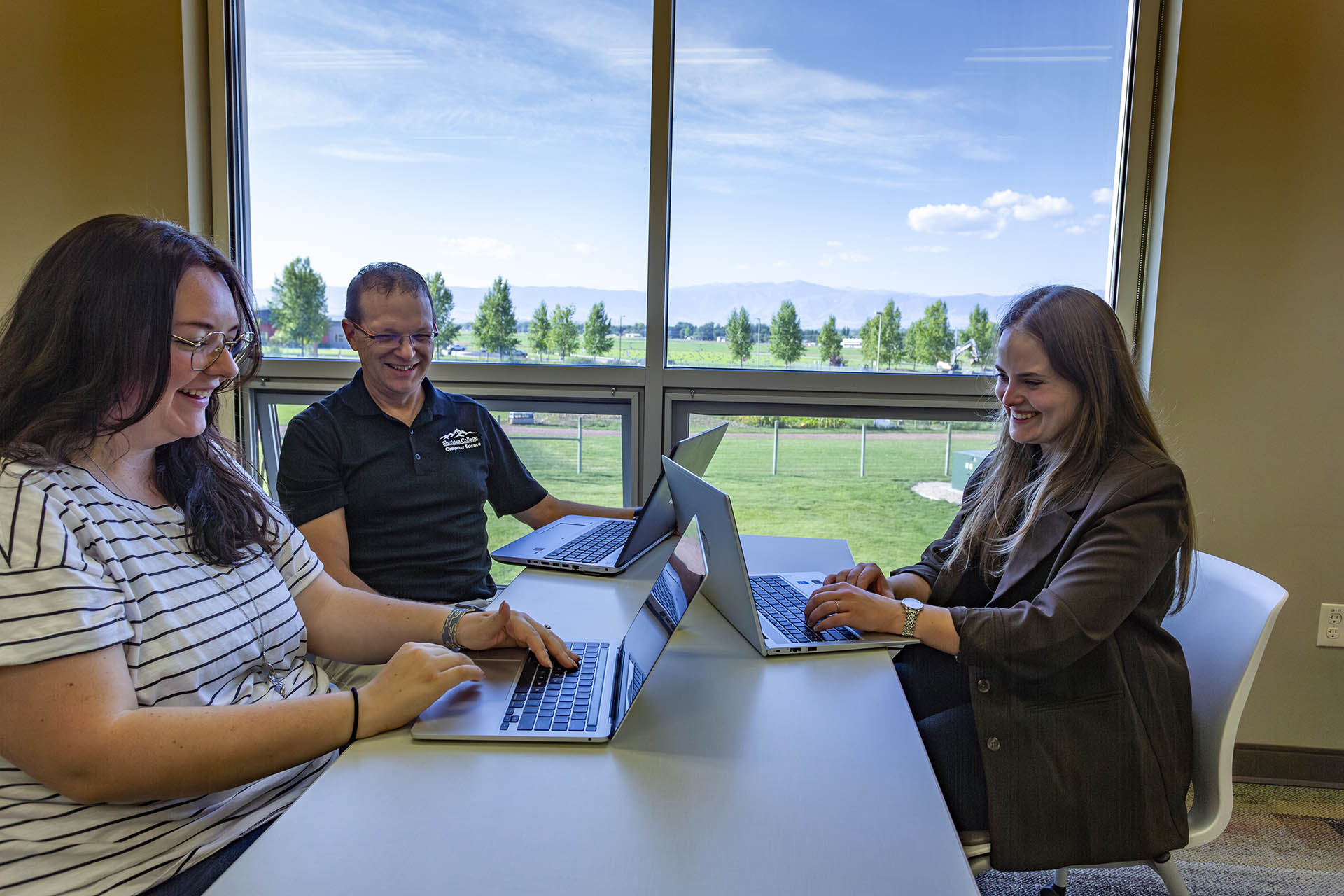 A Student works alongside two computer science instructors with a view of the Bighorn Mountains out the window of the classroom.
