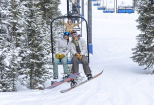 snowboarding at antelope butte, two female students smiling and riding the chairlift with one behind them also smiling and putting her hands up in the air excited