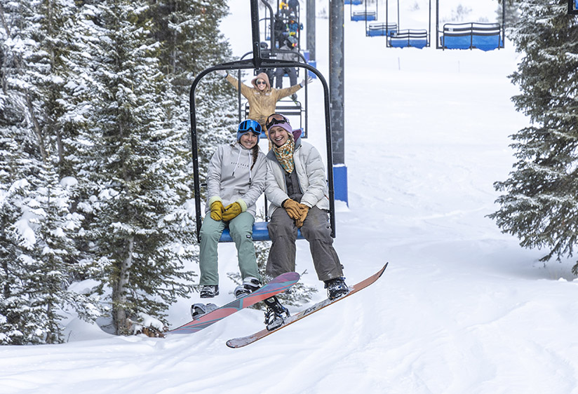 snowboarding at antelope butte, two female students smiling and riding the chairlift with one behind them also smiling and putting her hands up in the air excited