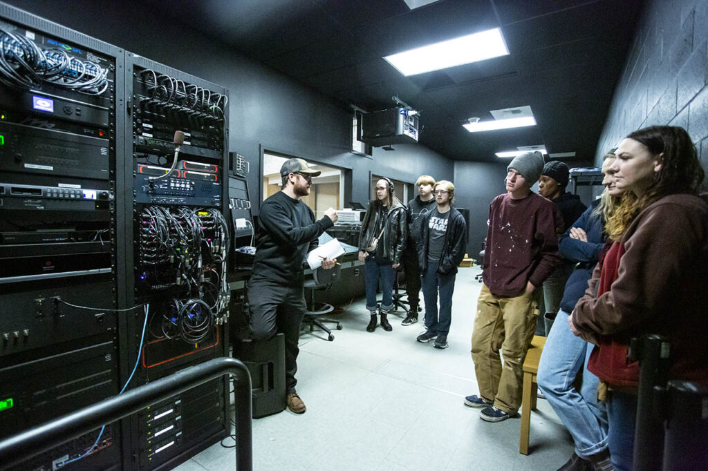 Students and technical director inside the booth in Kinnison Hall with a bunch of routing wires and patch cables visible in a tall rack tower.