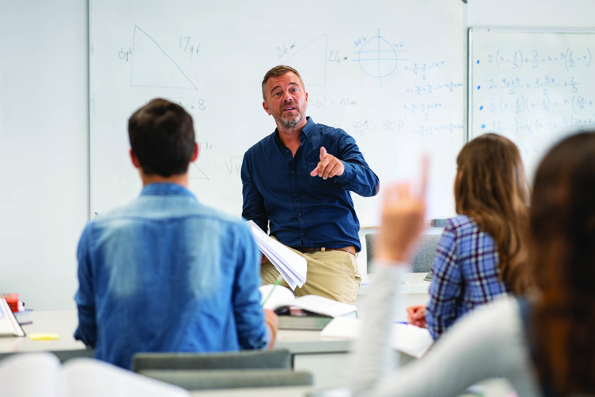 Professor pointing at high school student with hand raised in classroom. Student raising a hand with a question for the teacher. Lecturer teaching in class while girl have a question to do during a math lesson.
