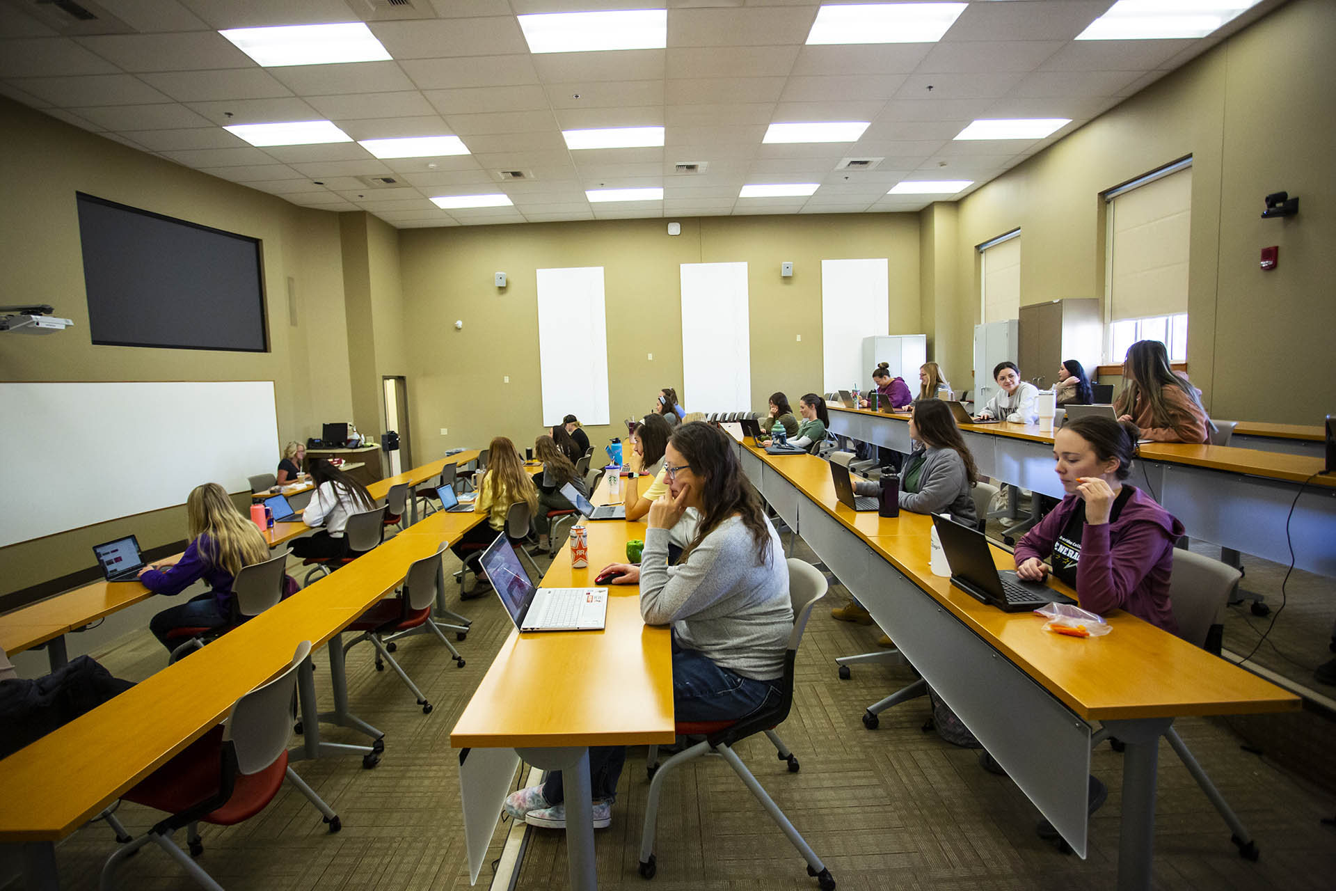 Students in a tiered classroom with several rows of desks, working on their laptops