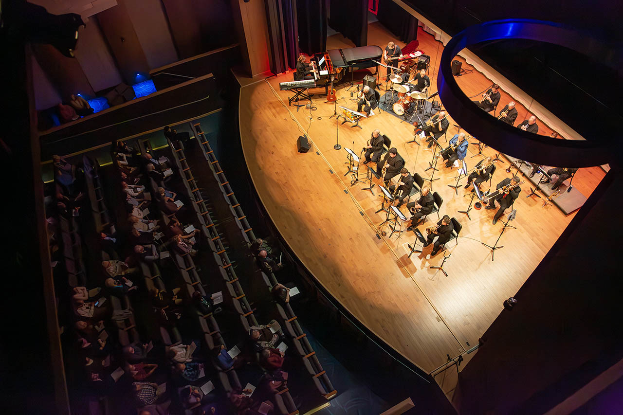 Photo taken from above the Bighorn Jazz Orchestra during a performance at Kinnison Hall looking down at the stage full of brass musicians, a piano, drumset and guitars.