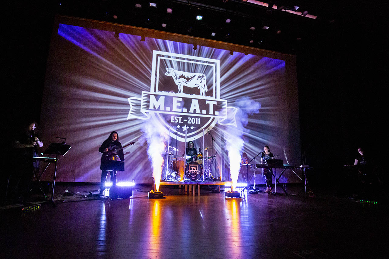Musicians on stage with the logo for Music Ensemble of Audio Technology or MEAT on the screen behind them with smoke machines shooting up on either side of the drums and guitar, wind instrument and multiple keyboards on stage.