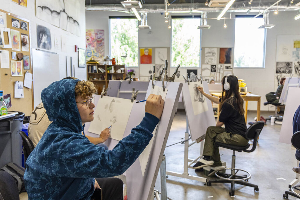 Art students working on drawings on easels throughout a brightly lit and colorful classroom.