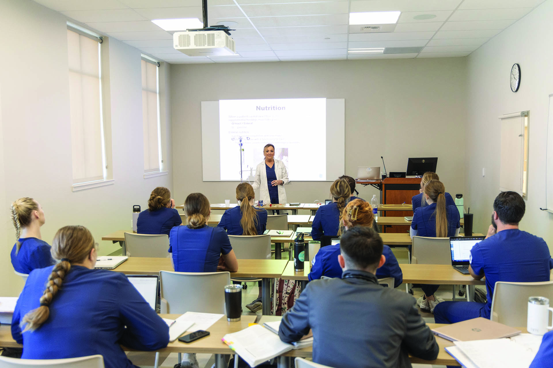 Image of an instructor in teaching in a brightly lit classroom with a presentation on the projector and room full of students in blue nursing scrubs