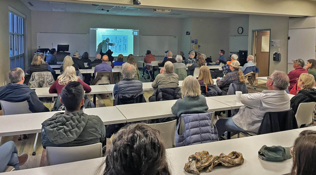 Photo of a lecturer pointing at a projection at the front of the classroom full of attendees.