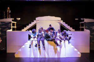 Actors on a theater set, crowded together and sitting at the top of some stairs during a production of The Snow