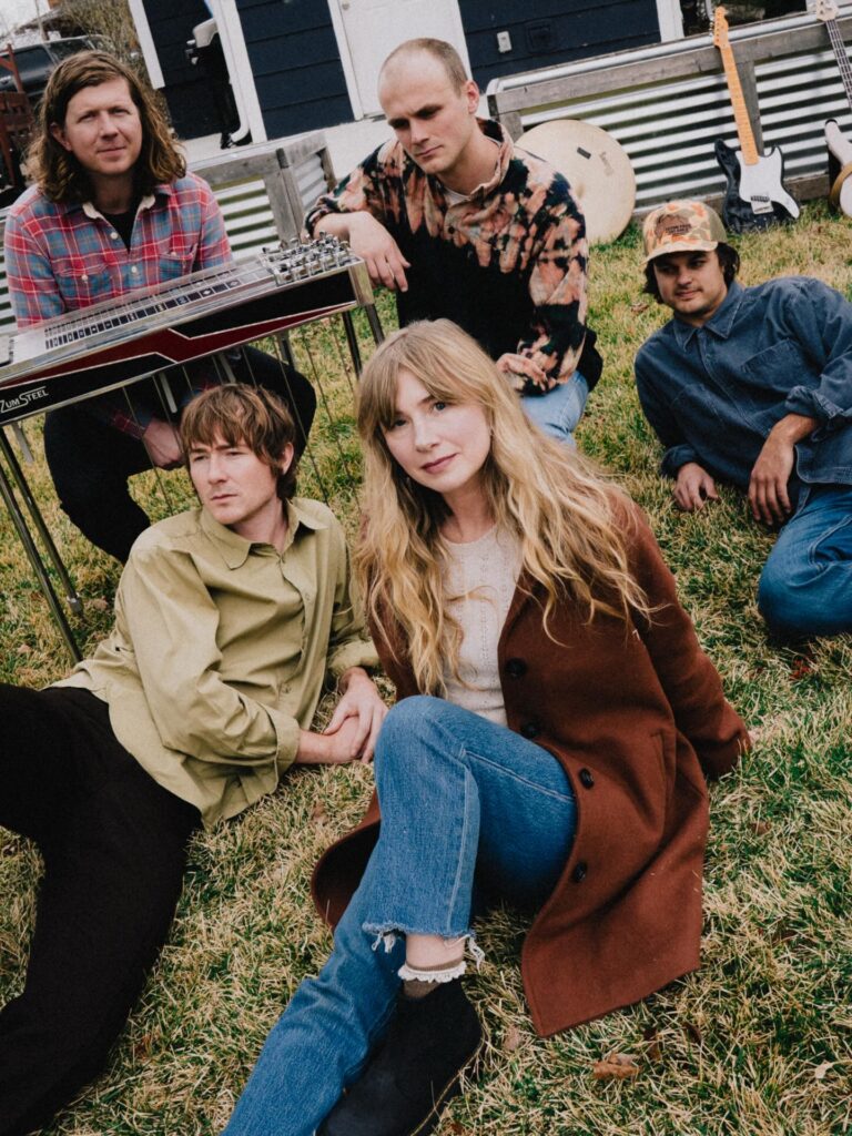 Photo of five band members sitting in the grass with a pedal steel