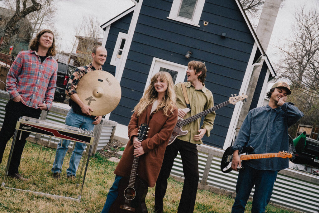 Abby Webster and her imaginary band standing with their instruments outside a blue house