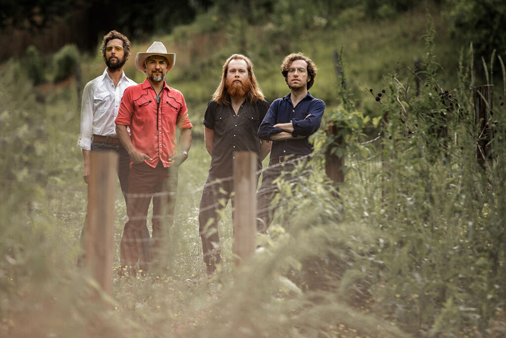 photo of the band outside behind a fence with green grass, western scene