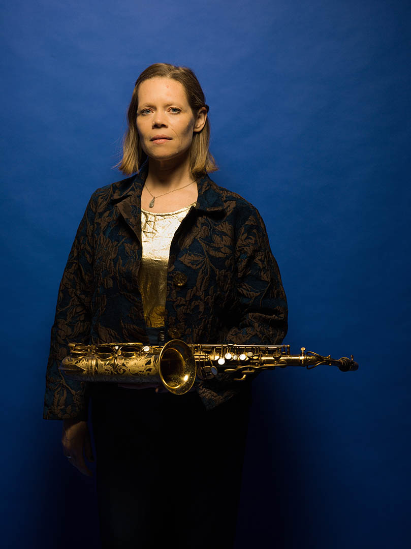 Caroline Davis standing in front of a blue curtain with her saxophone.